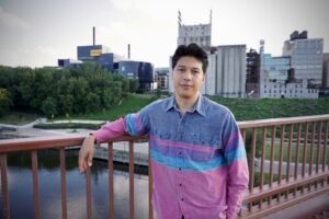 A person with light skin tone and black hair smiles softly wearing a pink, purple, and blue denim button-up shirt on a bridge in front of a city skyline.