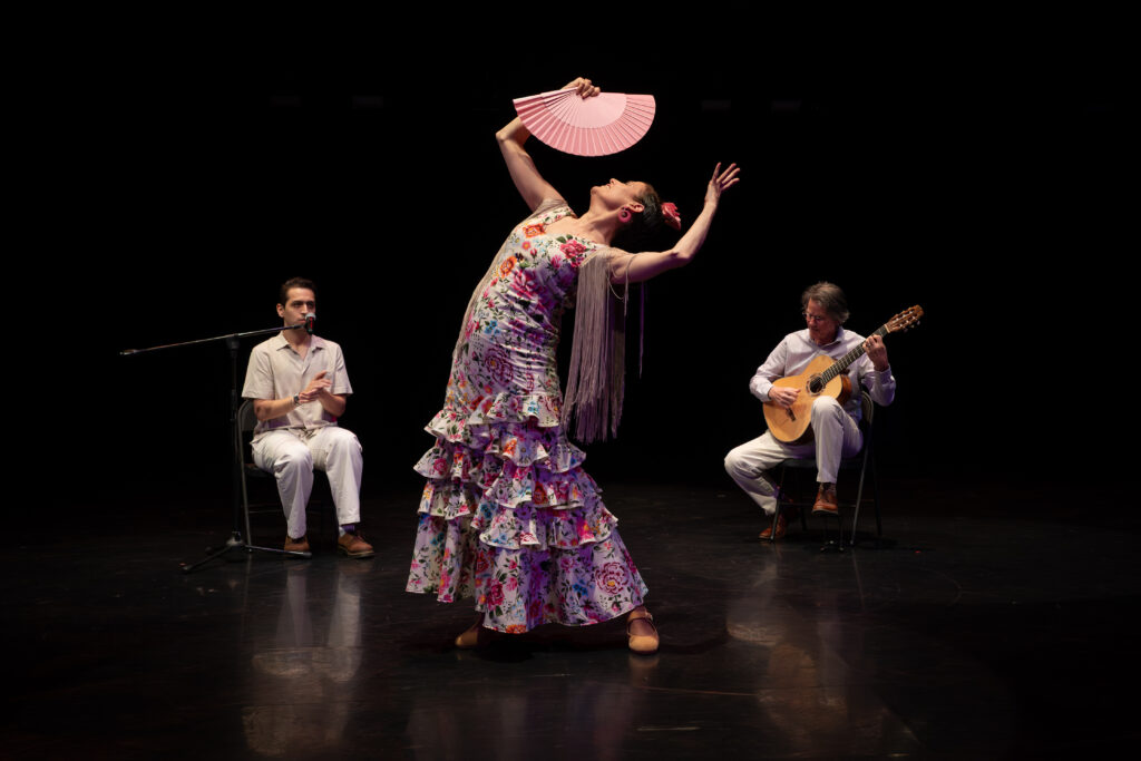 A person wearing a long tiered floral dress standing onstage with their back arched and arms above their head, holding a fan, while two seated musicians wearing white perform behind.
