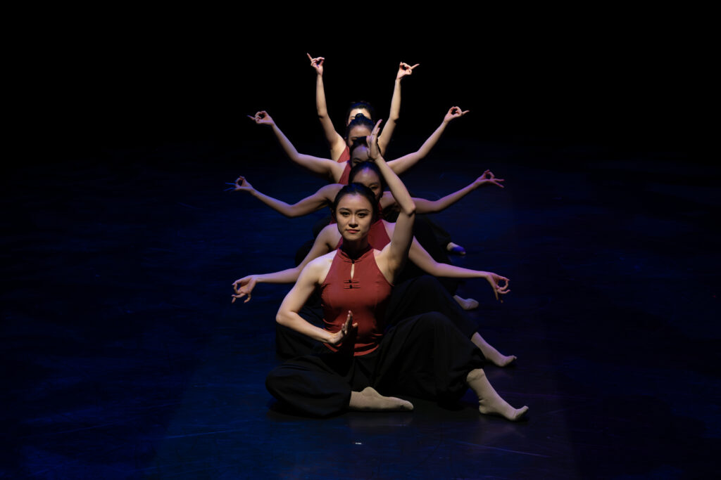 A person sitting on the ground with their arms outstretched in a dance performance, with four people sitting behind them with their arms in a similar outstretched pattern.