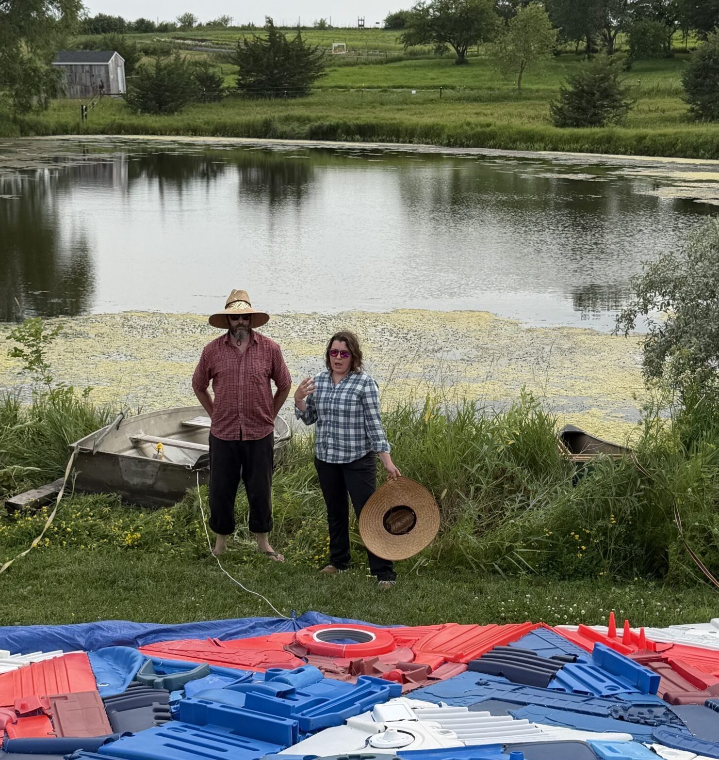 Two people standing by a pond with farmland in the background gesturing and talking