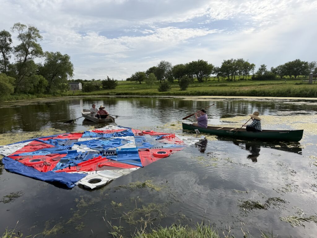 People in a paddle boat and canoe work together to pull a floating quilt-like sculpture on to a pond