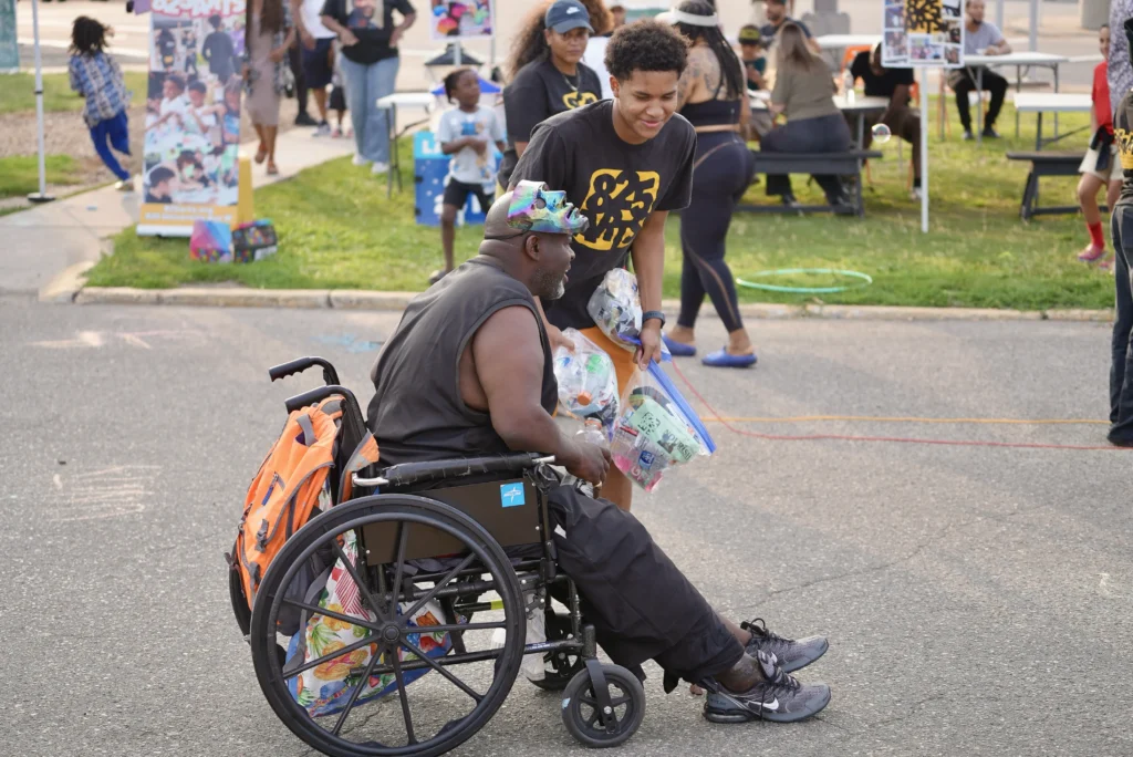 A person with dark skin wearing an 825 Arts t-shirt hands a community care package to a person in dark skin using a wheelchair.