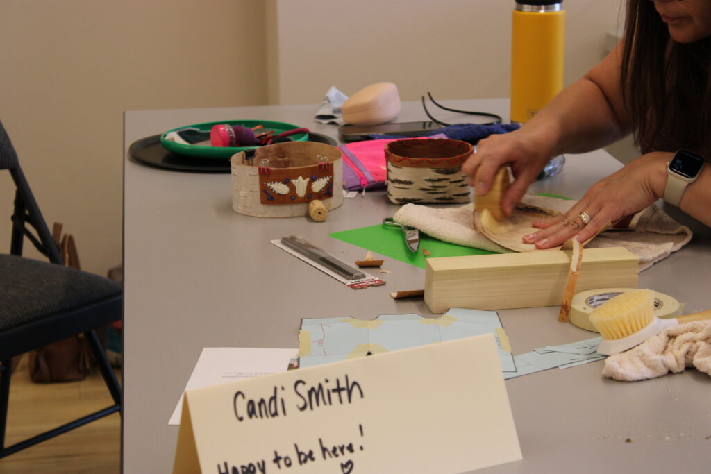 A person with light skin preparing bark for a basket during a workshop. Materials are spread across the table. A sign in the foreground reads, "Candi Smith. Happy to be here!"
