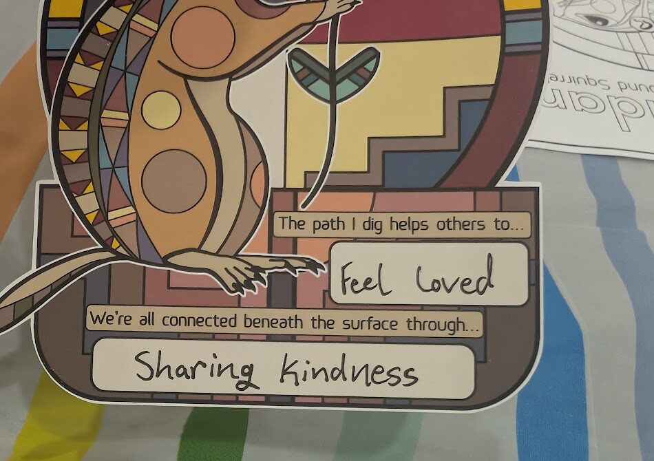 A closeup of a sticker with fill-in-the-blanks, reading: The path I dig helps others to... feel loved. And: We're all connected beneath the surface through... sharing kindness.