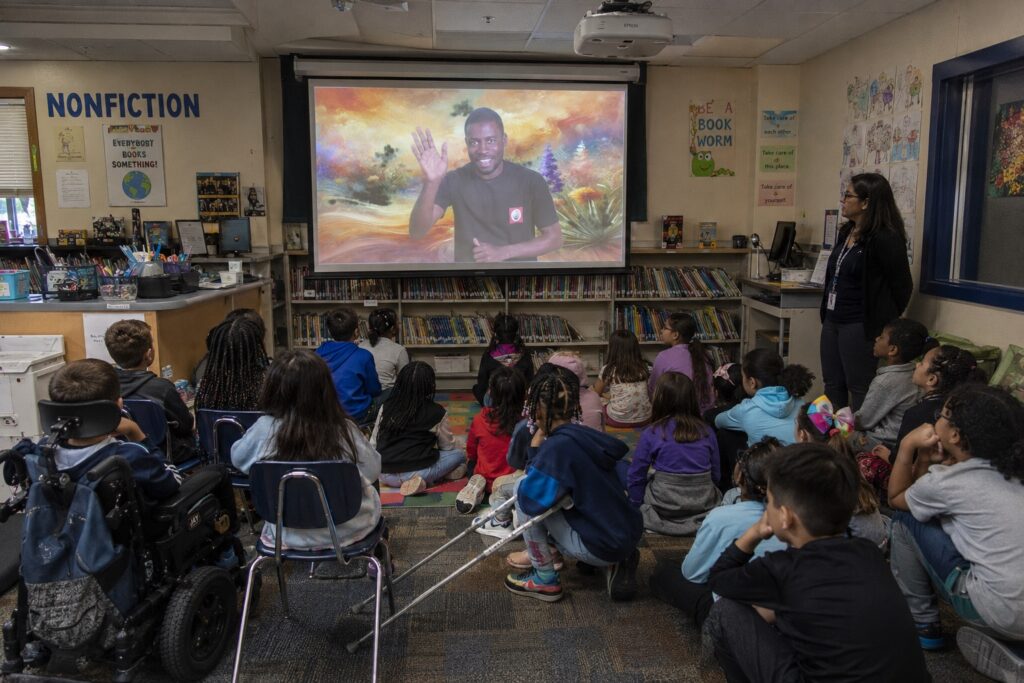 A second grade classroom with around two dozen young students and an adult teacher, sitting on the carpet and facing a projection screen featuring the sign language performer from "The Art of Accessible Sound."