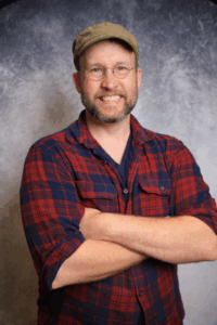 A headshot of a smiling person with light skin with brown facial hair, wearing a red and blue plaid shirt, small circular glasses, and a tan cap.
