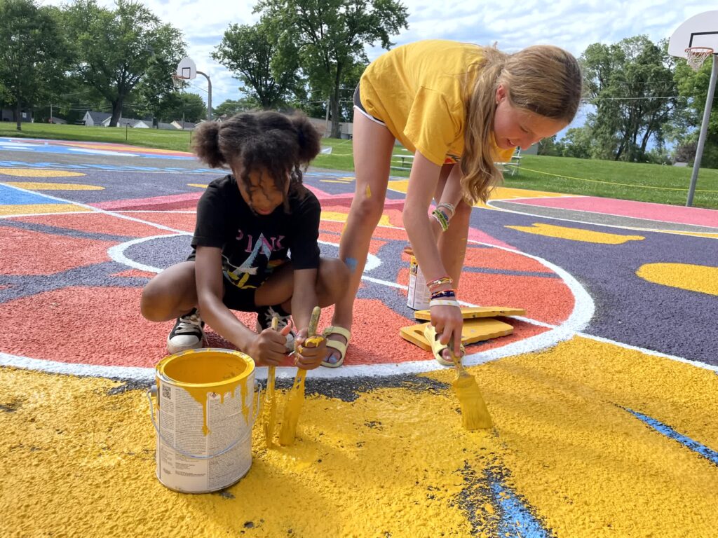 Two young people paint a basketball court mural.