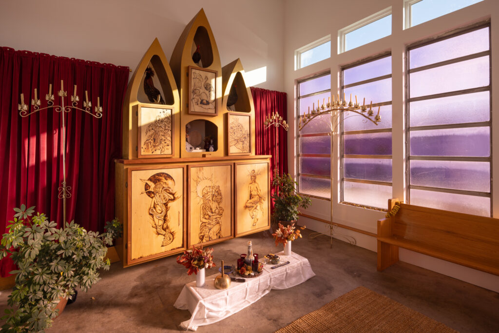 A warmly lit one-room chapel featuring a wooden pew, red curtains, intricate tall candle stands, a low altar covered in flowers and incense, and potted plants. In the center is a large wooden structure featuring engraved images of mythological creatures and three taxidermied chickens in alcoves.