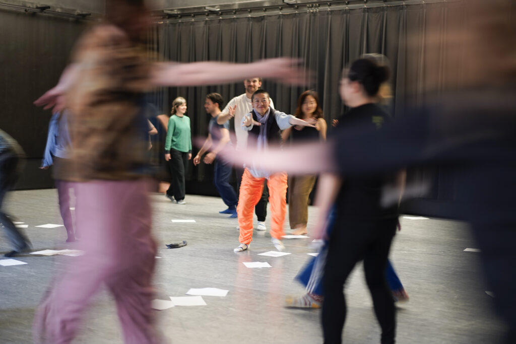 A person in orange pants stretches their hand toward the camera as blurred people move around the room.