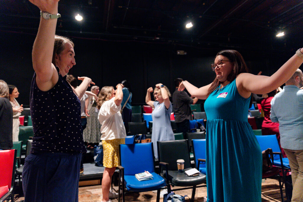 Two people face each other, grinning mid-stretch, as a room full of people moves together in a black box theater.